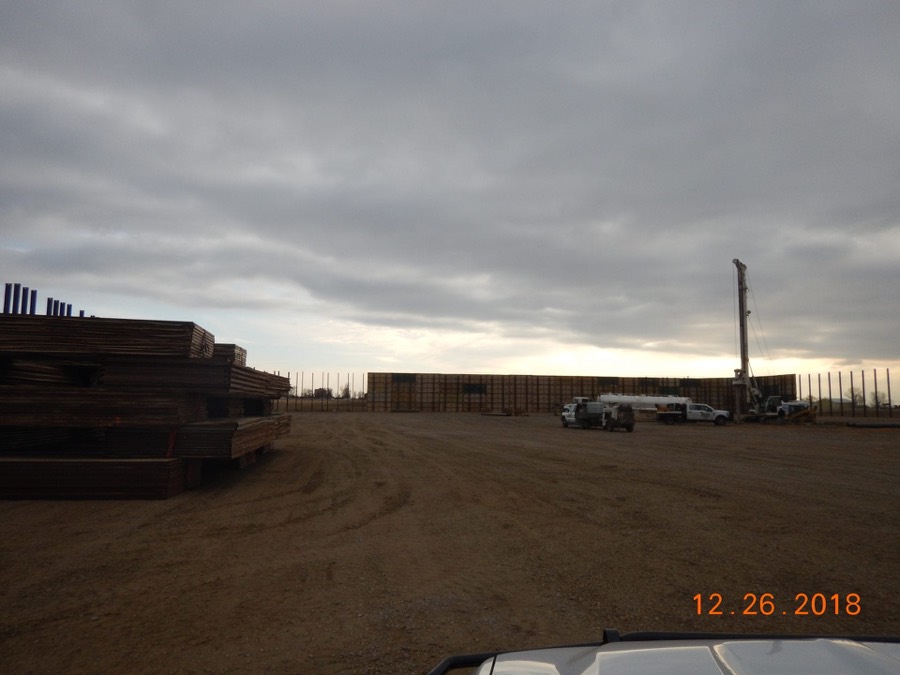 A drilling rig and stacked rig mats behind a sound wall on an overcast winter morning
