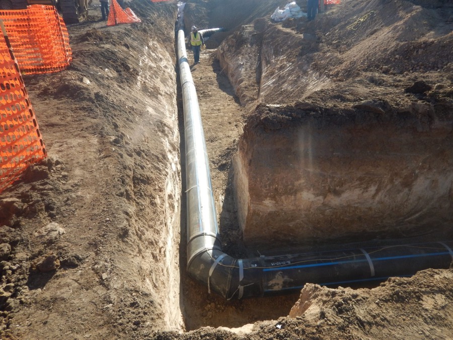 A large diameter steel water line being set into an open trench by a crew in high visibility gear
